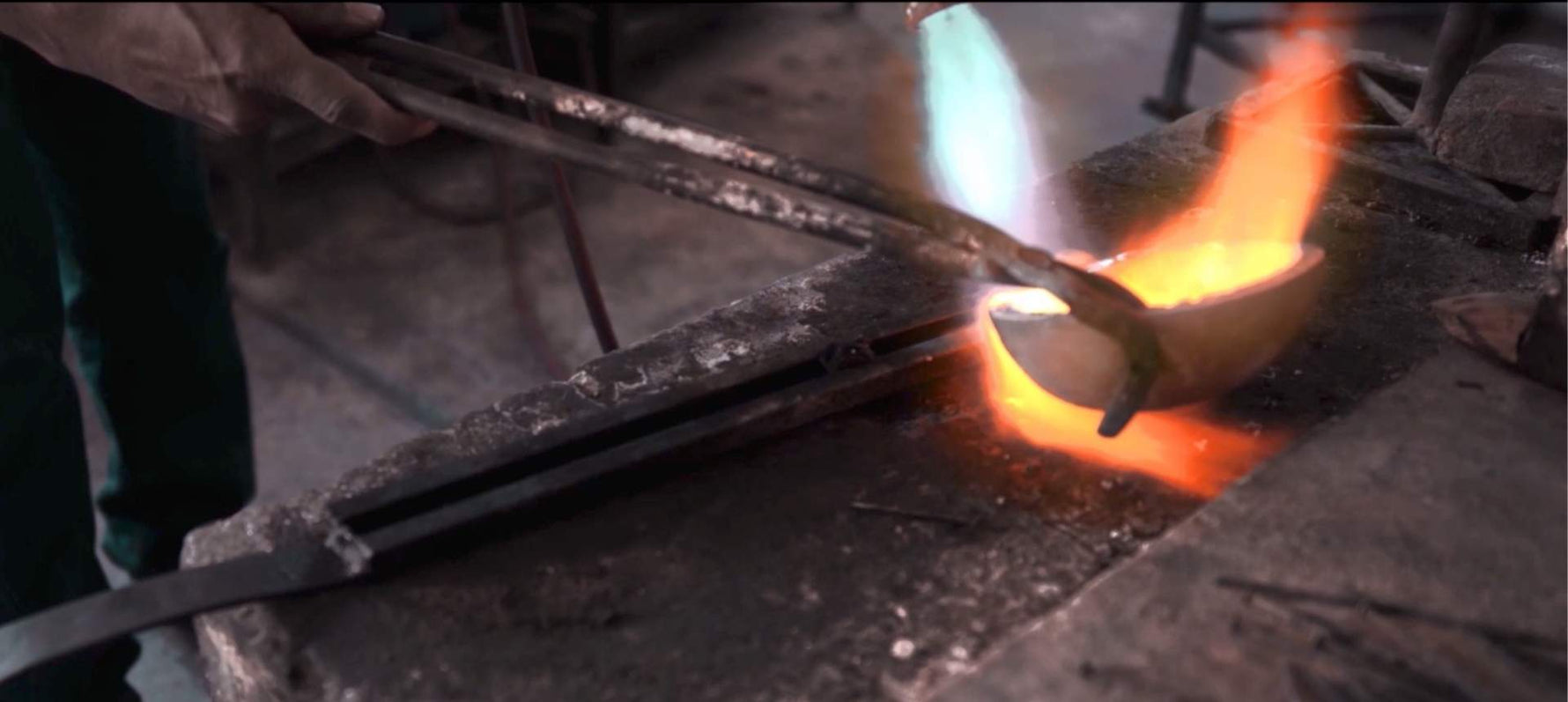 Close-up view of silversmith's hands using traditional tongs to tilt a glowing crucible over intense blue and orange flames, melting solid silver in a Taxco artisan workshop.