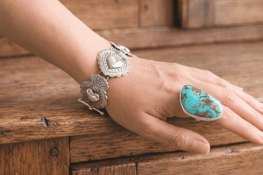 Handcrafted silver Sacred Heart bracelet and a bold turquoise statement ring on a woman's hand, resting on a rustic wooden cabinet to highlight artisan Taxco jewelry.