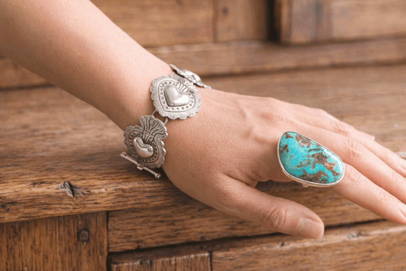Handcrafted silver Sacred Heart bracelet and a bold turquoise statement ring on a woman's hand, resting on a rustic wooden cabinet to highlight artisan Taxco jewelry.