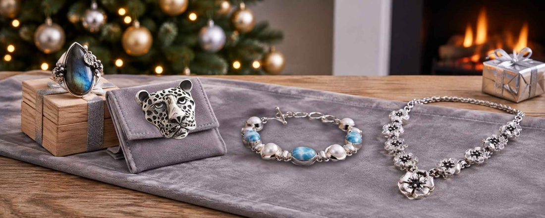 A festive holiday display featuring a labradorite ring, a leopard jewelry pouch, a larimar and pearl bracelet, and a silver flower necklace arranged on a grey velvet runner with Christmas decorations.