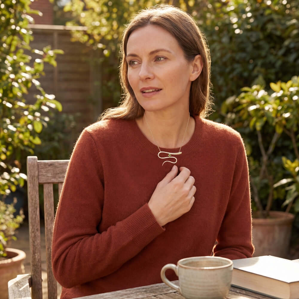 A woman in an orange sweater wearing a handmade silver wavy pendant with a green tourmaline gemstone while sitting in a sunlit garden.