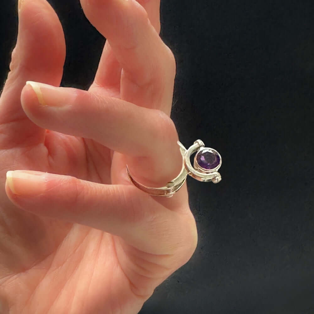 Hand wearing a silver ring with a purple gemstone against a dark background