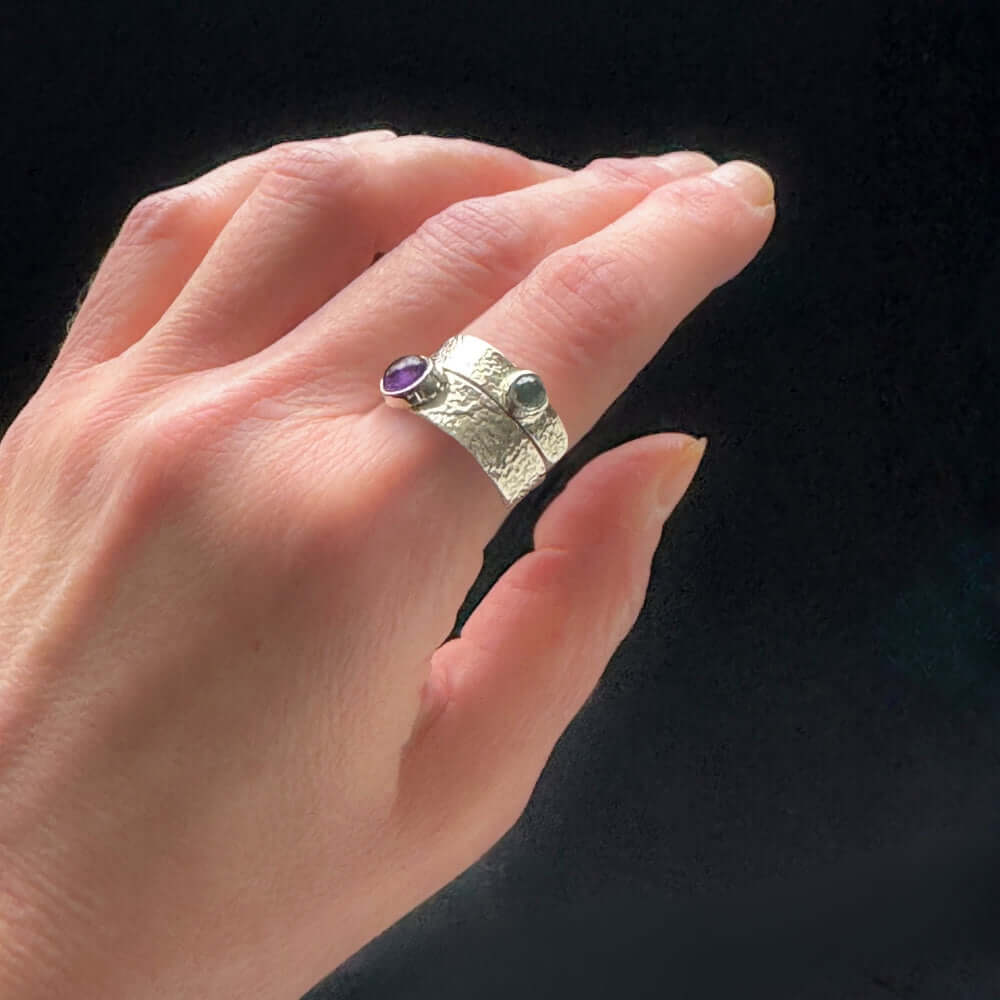 Hand wearing a silver ring with purple and light blue gemstones on a black background