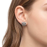A close-up of a woman's ear wearing an owl stud earring. The earring has a textured finish with blackened details in the owl's eyes and around its body.