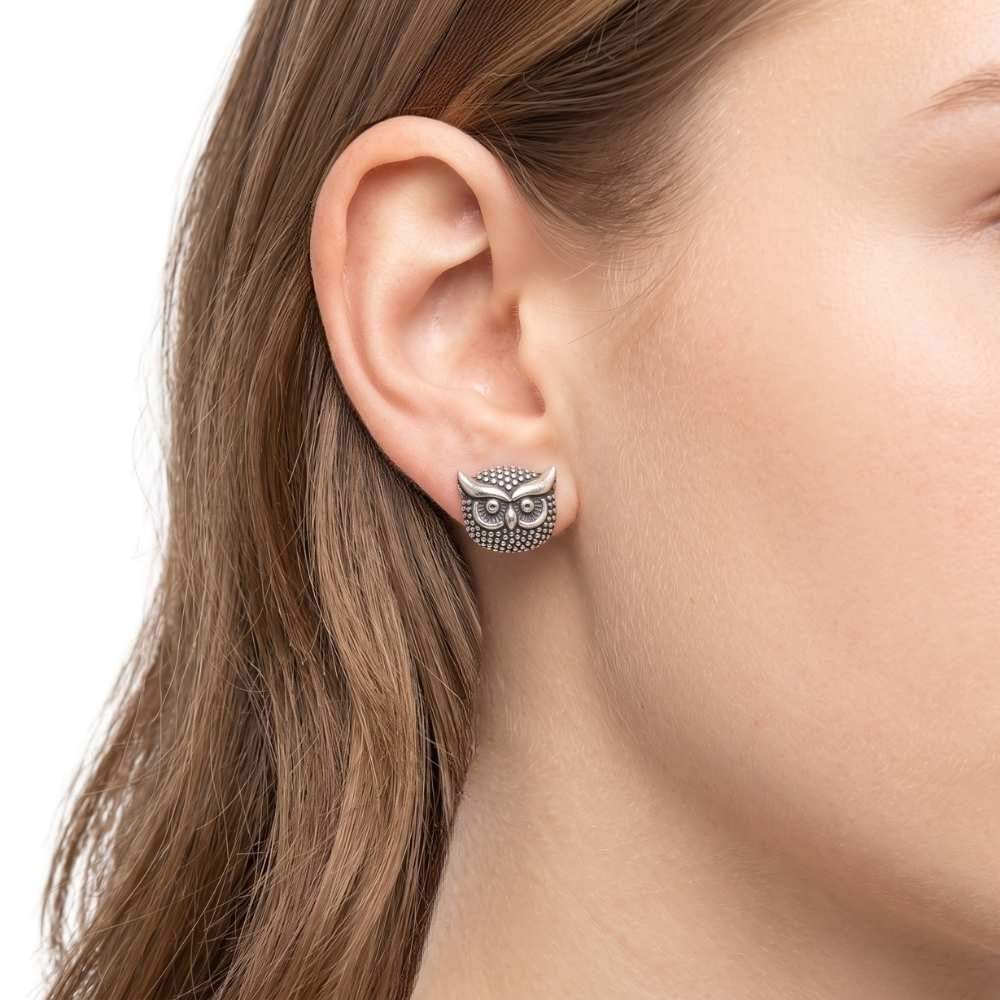 A close-up of a woman's ear wearing an owl stud earring. The earring has a textured finish with blackened details in the owl's eyes and around its body.