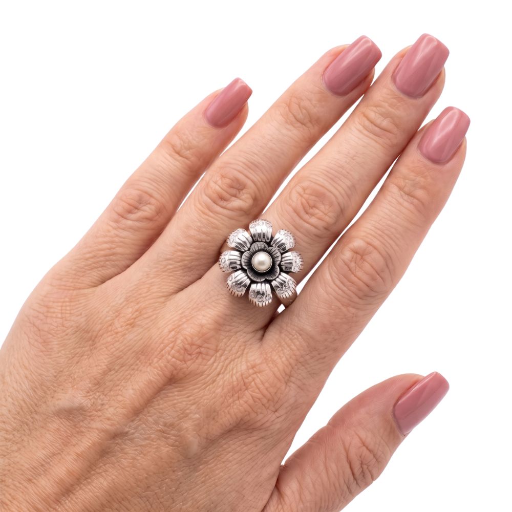 A silver-toned flower ring with a central pearl, worn on a hand with pink manicured nails against a white background.