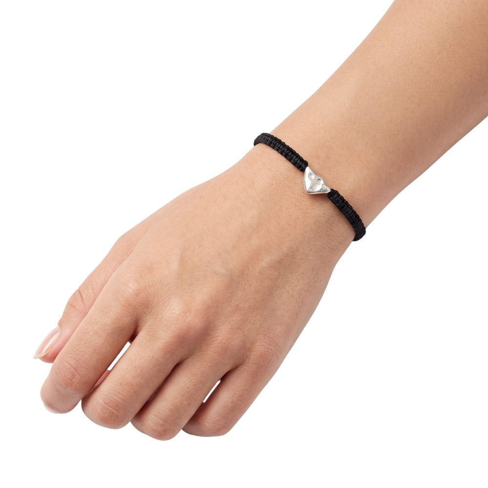 A close-up of a person's hand and wrist wearing a black braided cord bracelet with a small silver textured triangle, set against a plain white background.