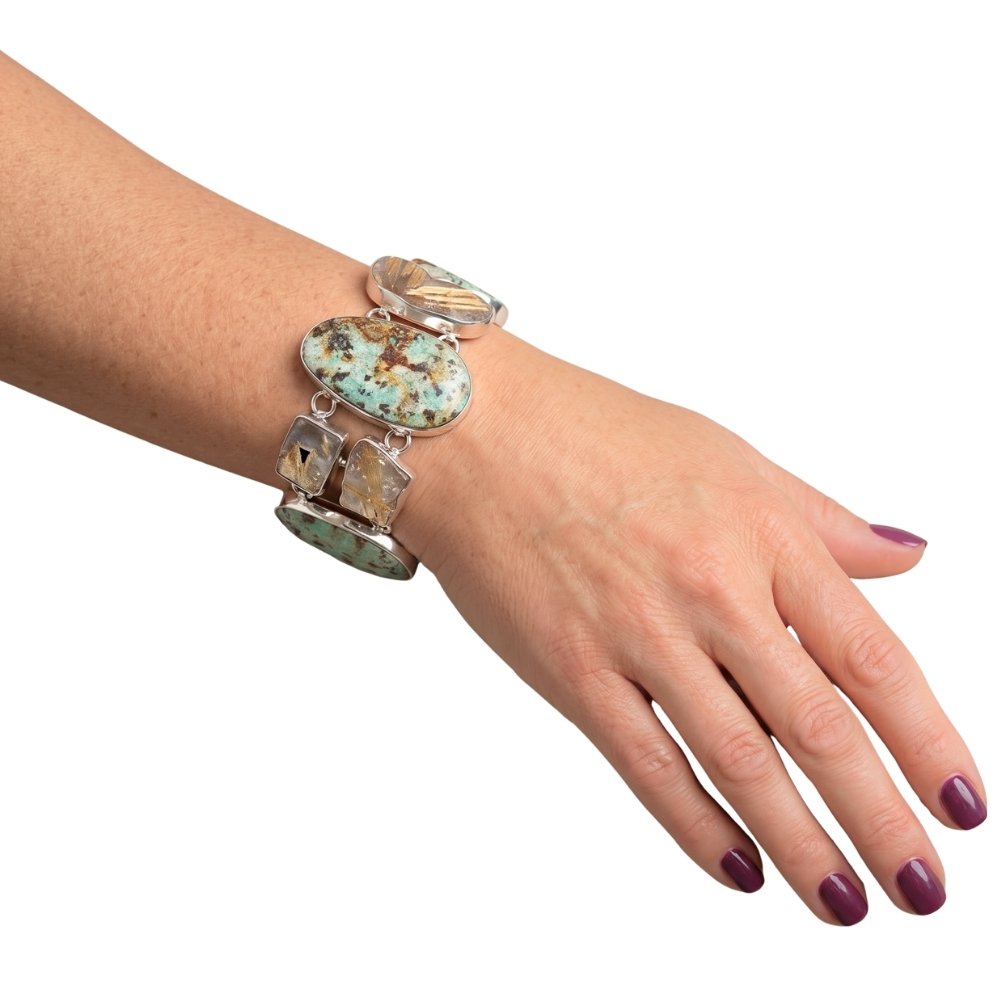 A close-up of a person's hand and wrist wearing a wide Taxco silver bracelet with green turquoise and rutilated quartz, set against a plain white background.