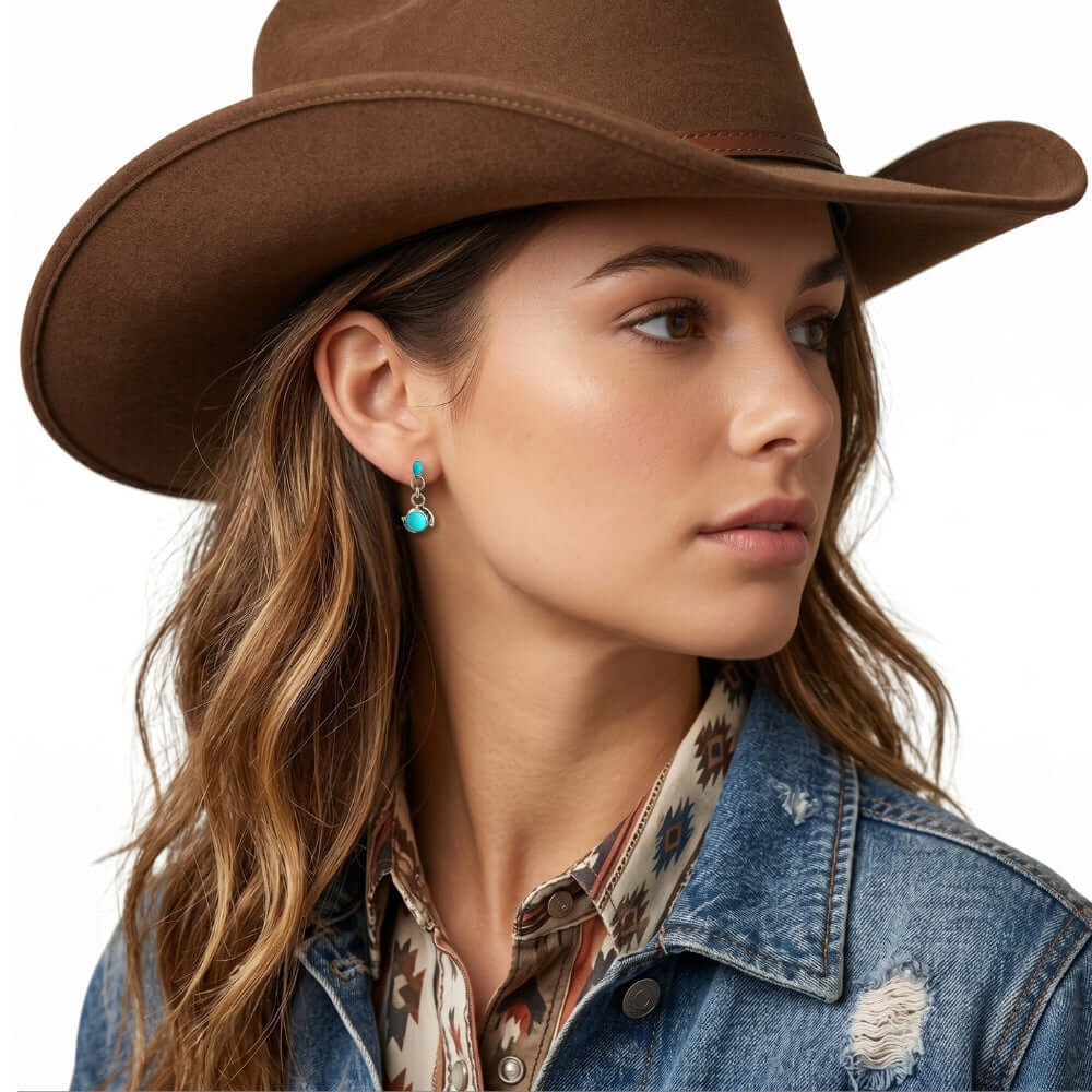 Woman wearing silver turquoise earrings, a brown cowboy hat, denim jacket, and patterned shirt on a white background