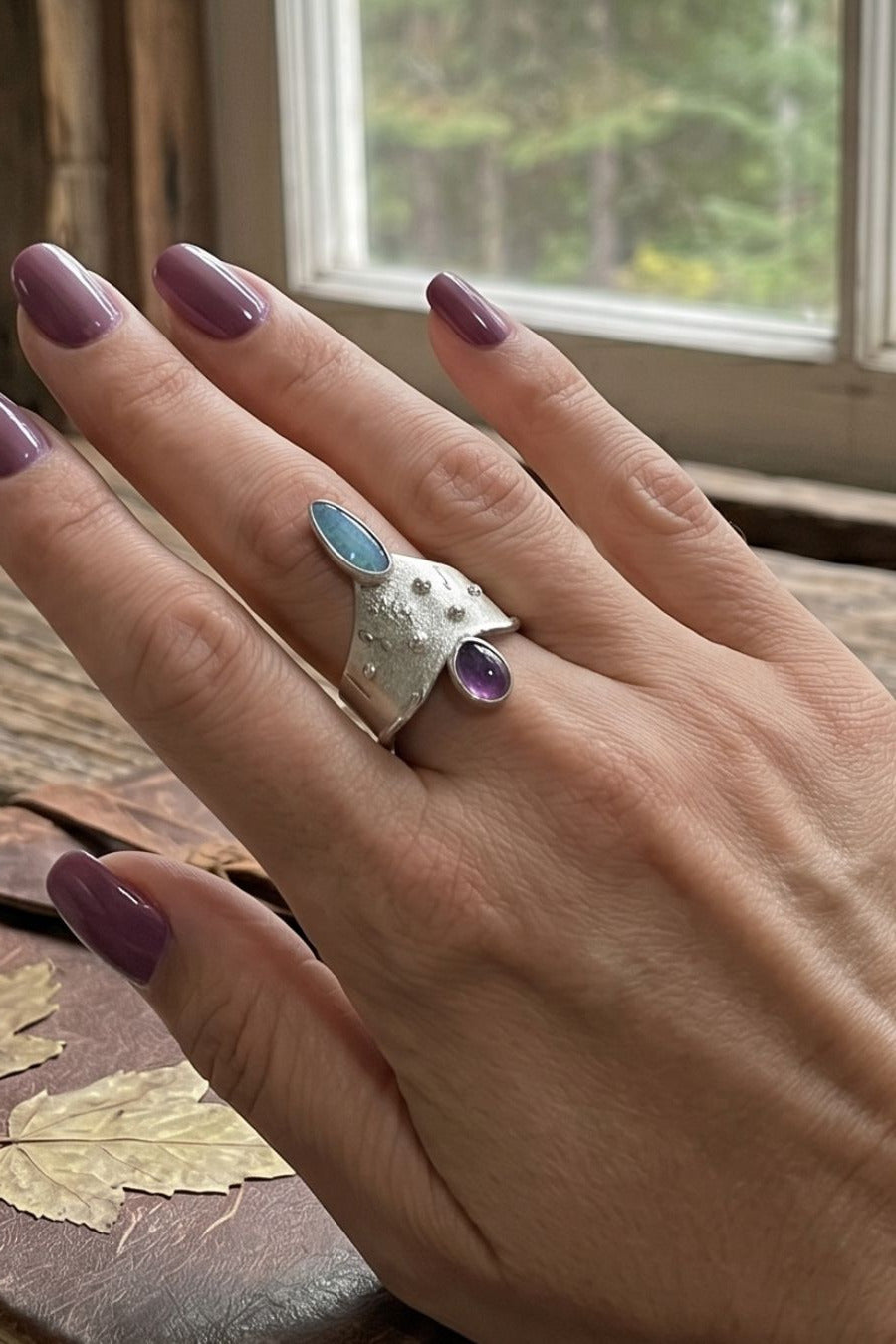 A hand wearing a unique Taxco silver ring featuring a textured band with opal and amethyst, set against a rustic window background.