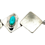A close-up of a silver-toned geometric link bracelet featuring a square link with a bezel-set oval turquoise stone and a textured square link beside it, on a white background.