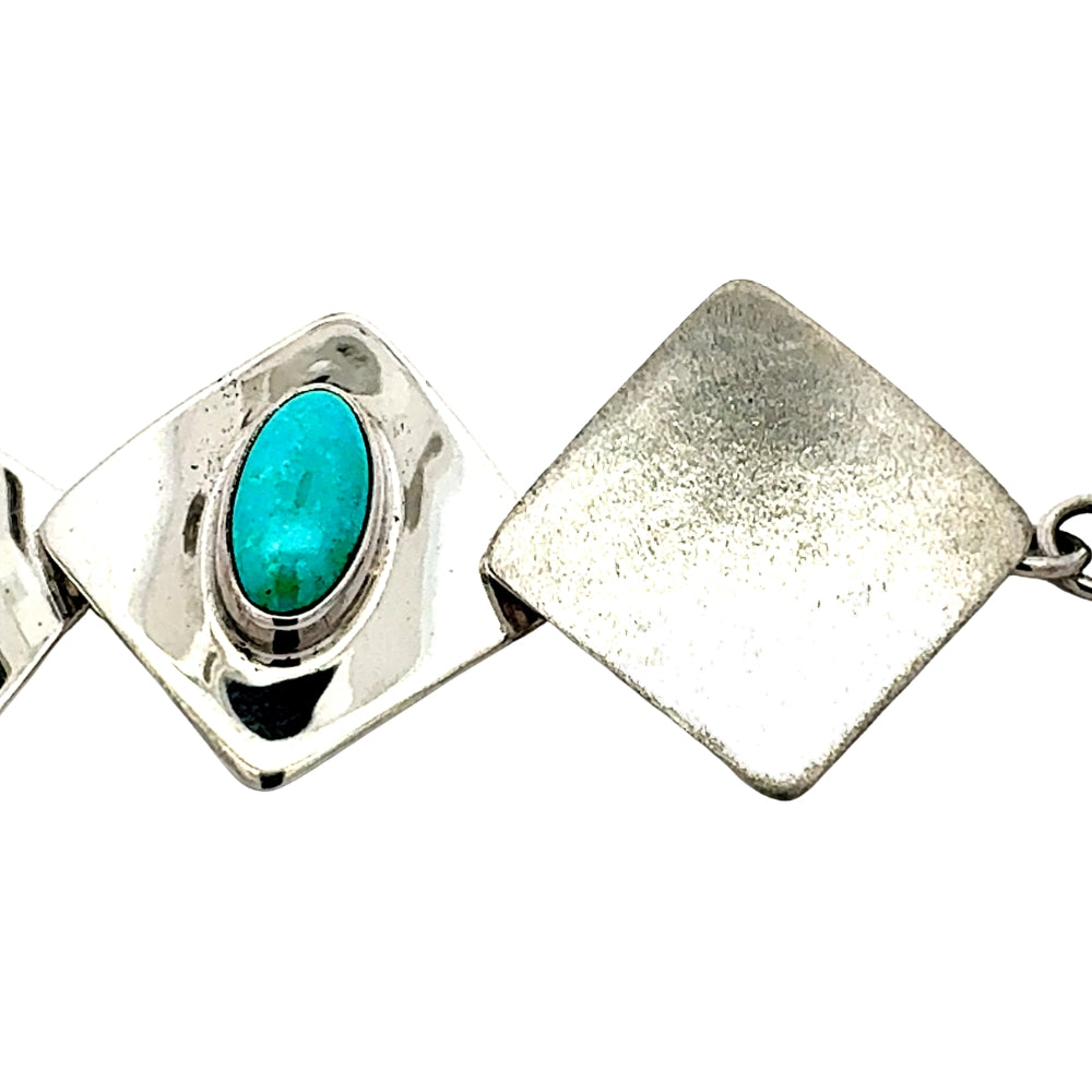 A close-up of a silver-toned geometric link bracelet featuring a square link with a bezel-set oval turquoise stone and a textured square link beside it, on a white background.