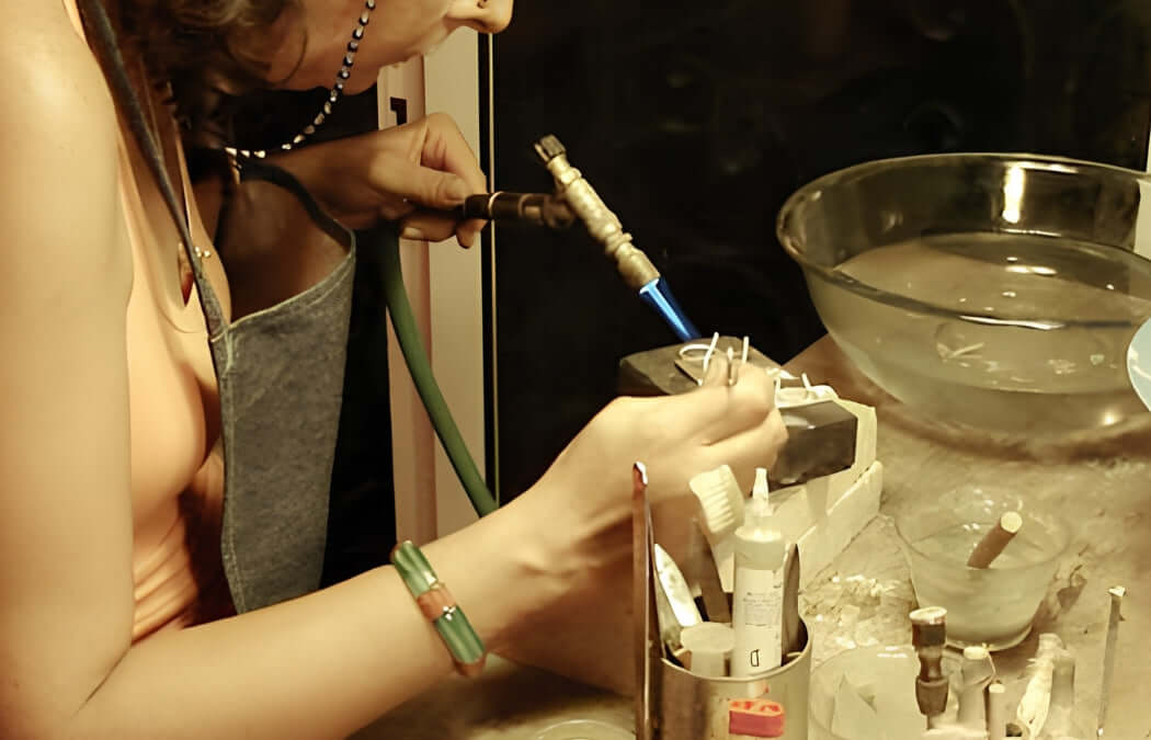 A close-up lifestyle shot of a jeweler using a torch with a blue flame to work on a piece of jewelry in a workshop setting. 